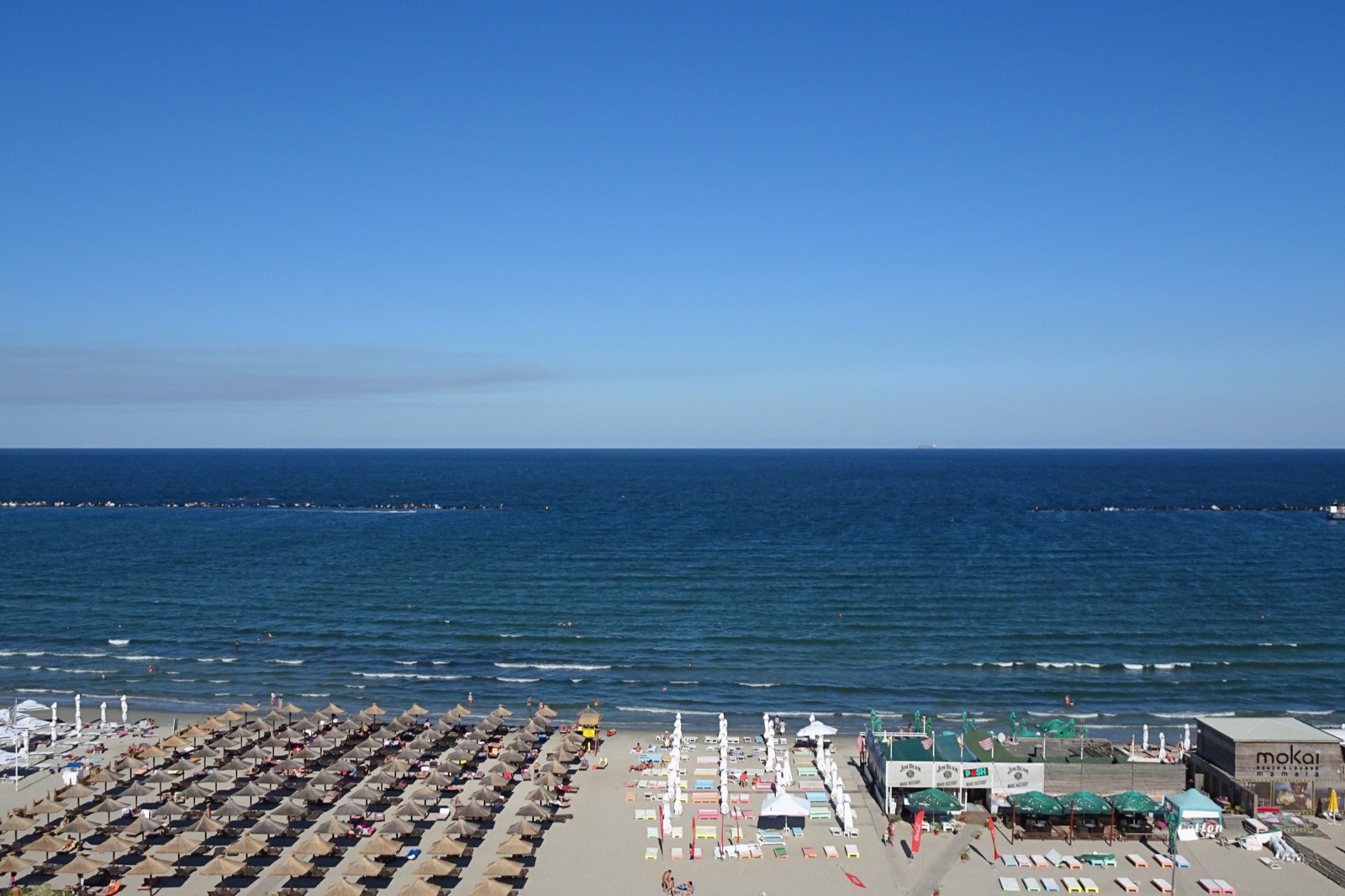Strohschirme und Liegen am Strand im Zentrum des Badeorts, dahinter das blaue Meer mit Wellenbrechern aus Stein