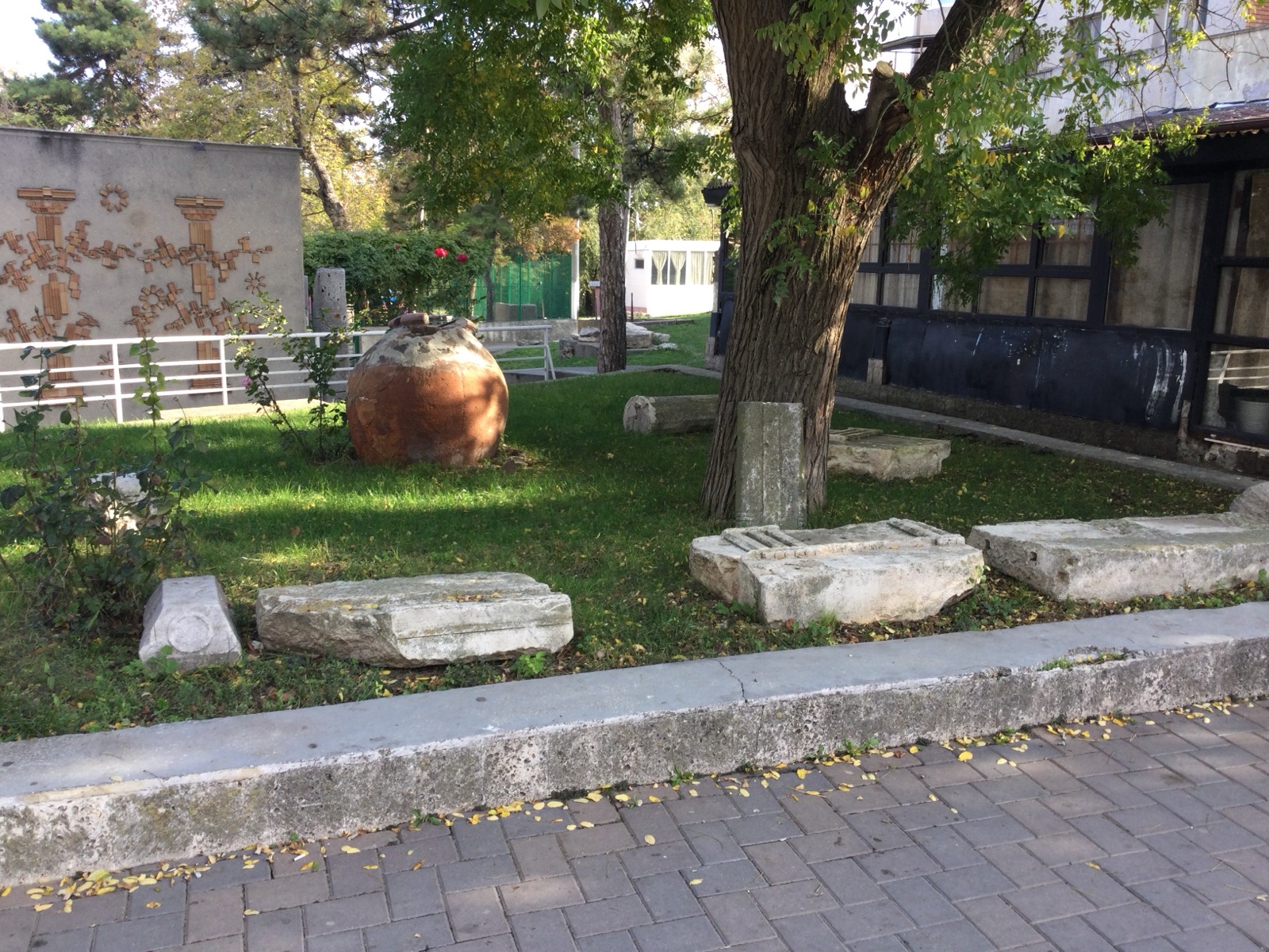Lapidariul din curtea muzeului, cu un vas mare de lut și blocuri de piatră antice așezate în iarbă